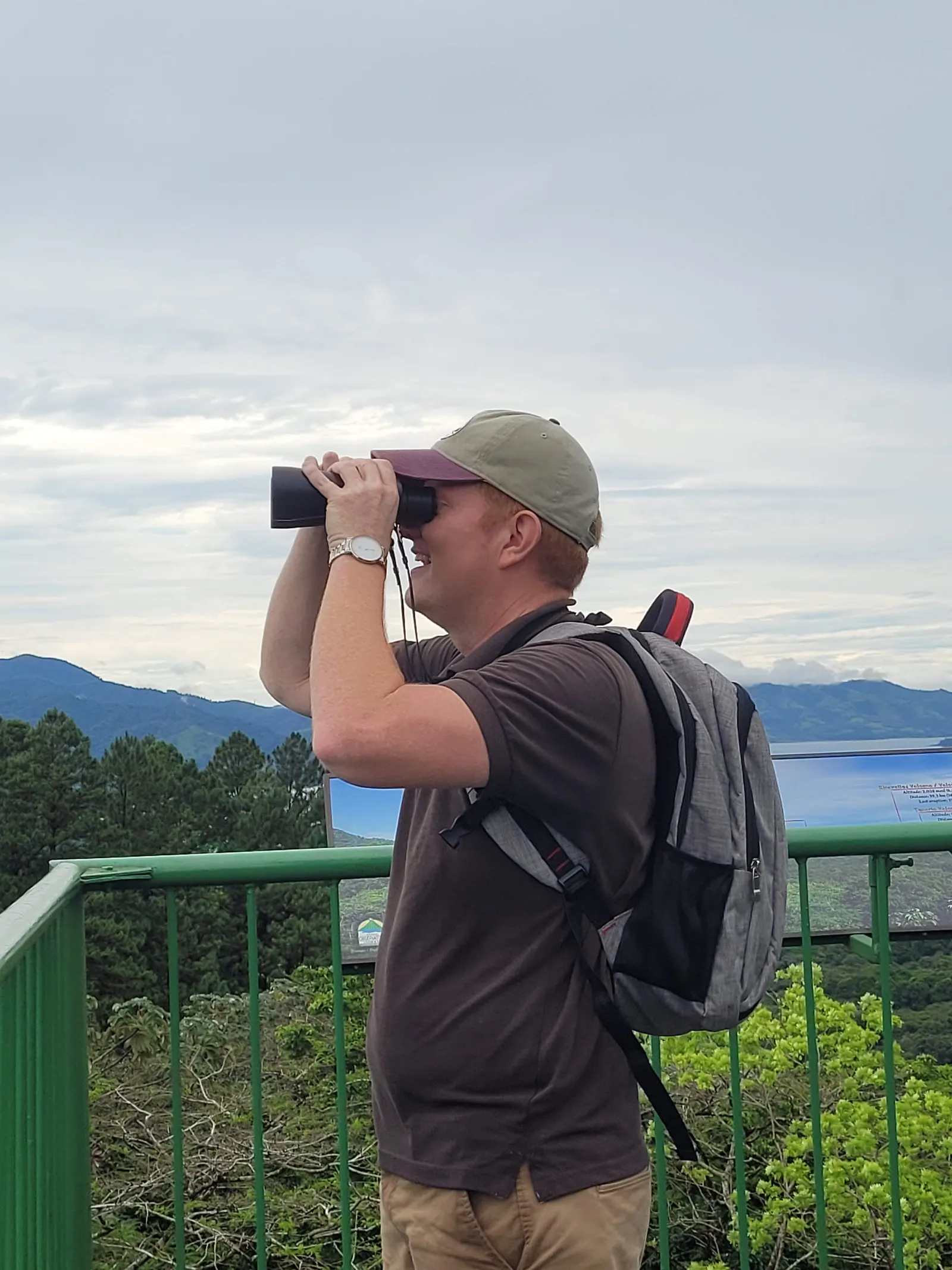 A man in a cap looks through binoculars from a railed lookout, green hills and a lake behind him.