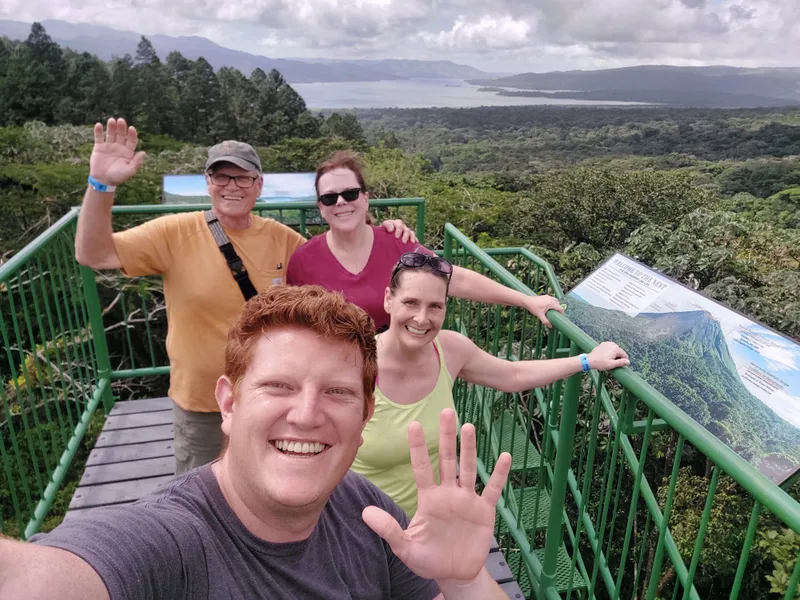 Four people at a green railing on a lookout, with forest and a lake spread out below.