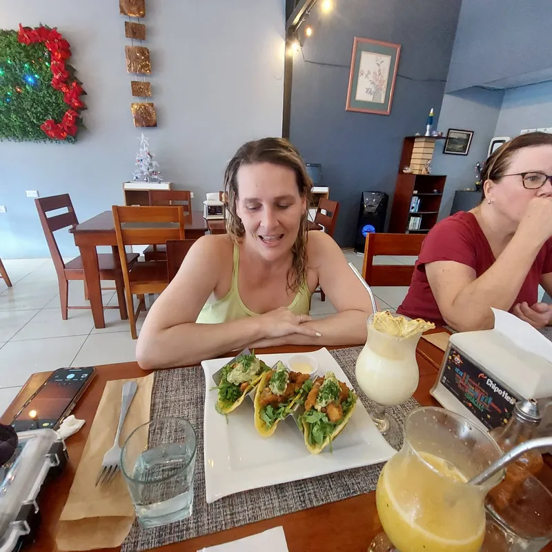 A woman sits at a restaurant table with a plate of tacos and two cold drinks in front of her.