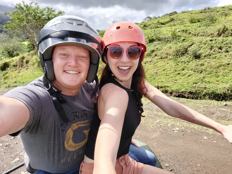 Two people in helmets take a selfie on a dirt road, with a steep green pasture behind them.