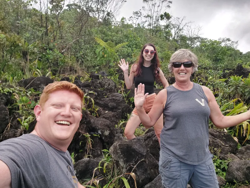 Three people stand among dark lava boulders, with forest growing up the slope behind them.