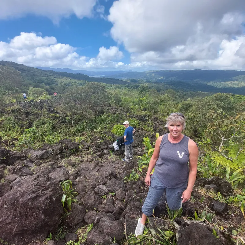 A woman stands on a field of dark boulders, with a wide valley and tall clouds behind her.