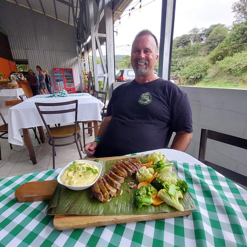 A man stands at a table set with grilled meat, mashed vegetables and salad served on a banana leaf.