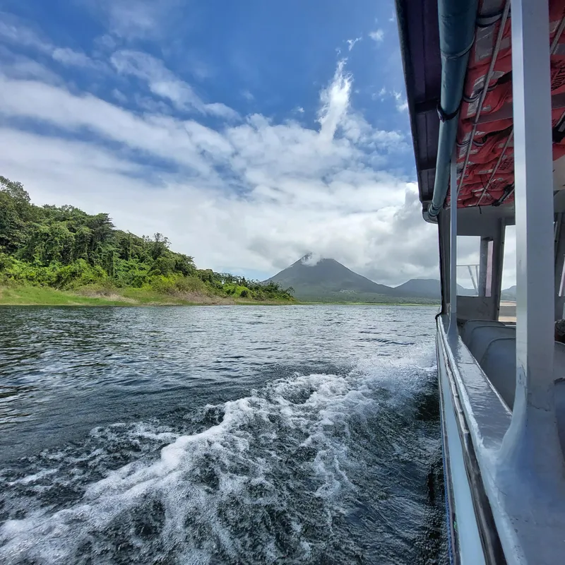 The wake of a boat on a lake, with a forested shore and a volcano in the distance.