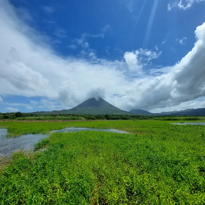 Flooded green pasture with water lying in patches and a volcano on the far side under heavy clouds.