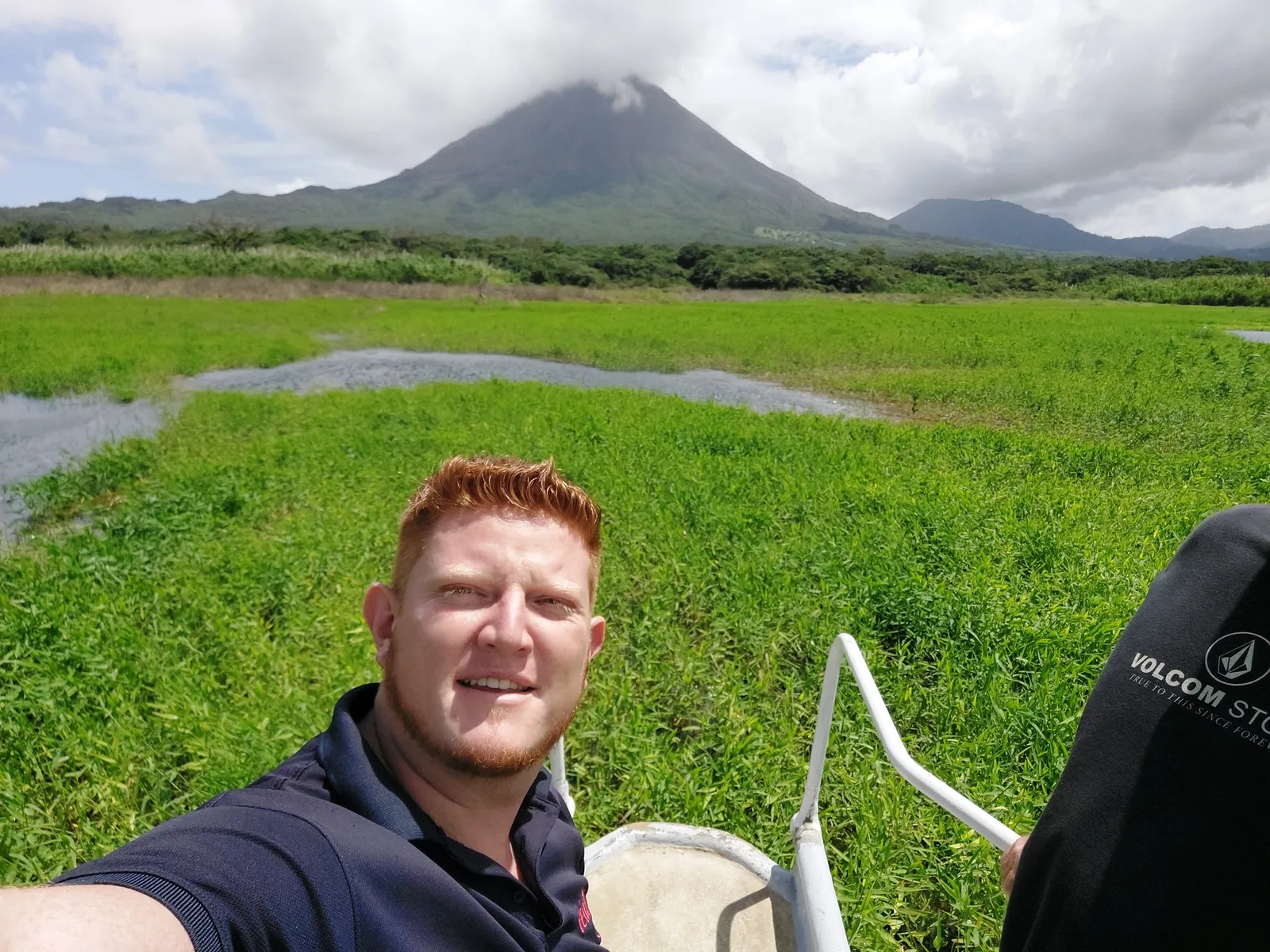 A man takes a selfie with wet green pasture behind him and a conical volcano in the distance.