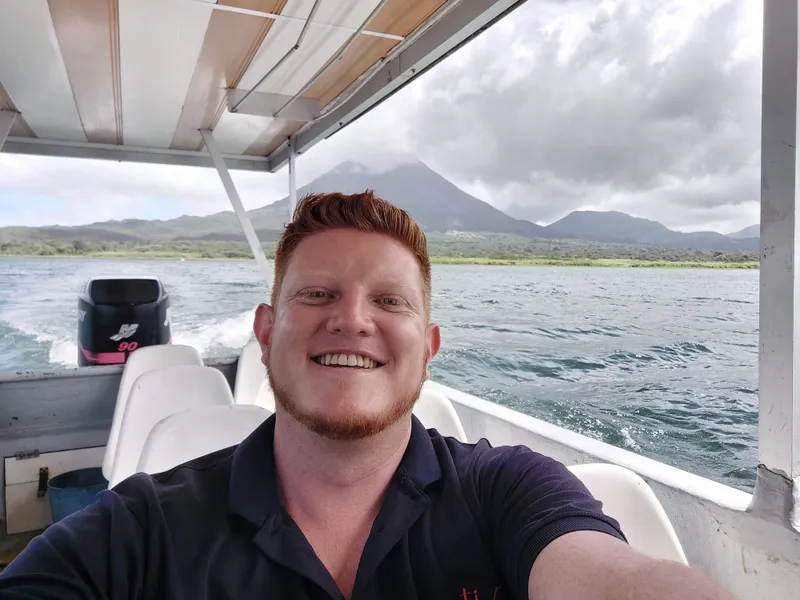 A man takes a selfie aboard a covered boat, with the lake and a volcano behind him.