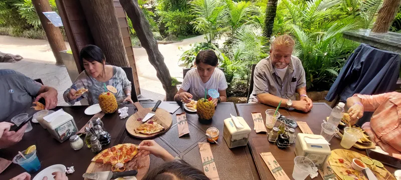 A table with pizza on a wooden board and a drink served in a hollowed pineapple, people eating around it in a garden.