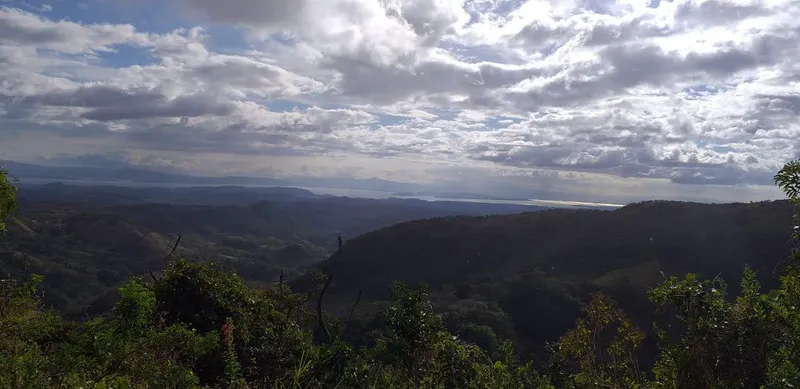 A dark forested valley seen from above, with a lake on the horizon and tall clouds over it.