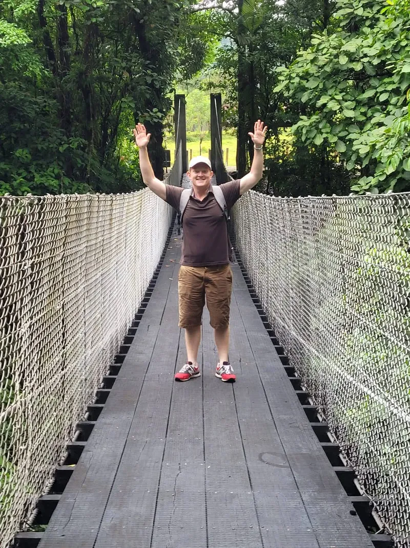 A man stands with both arms raised in the middle of a long suspension bridge in the forest.