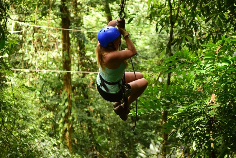 A woman hangs from a zip line cable with the forest behind her.