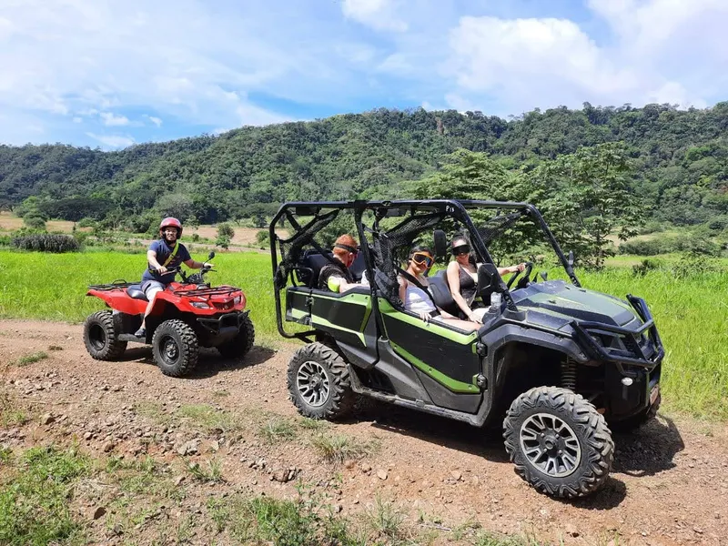 A quad and a side by side buggy stopped on a dirt track, green hills behind.
