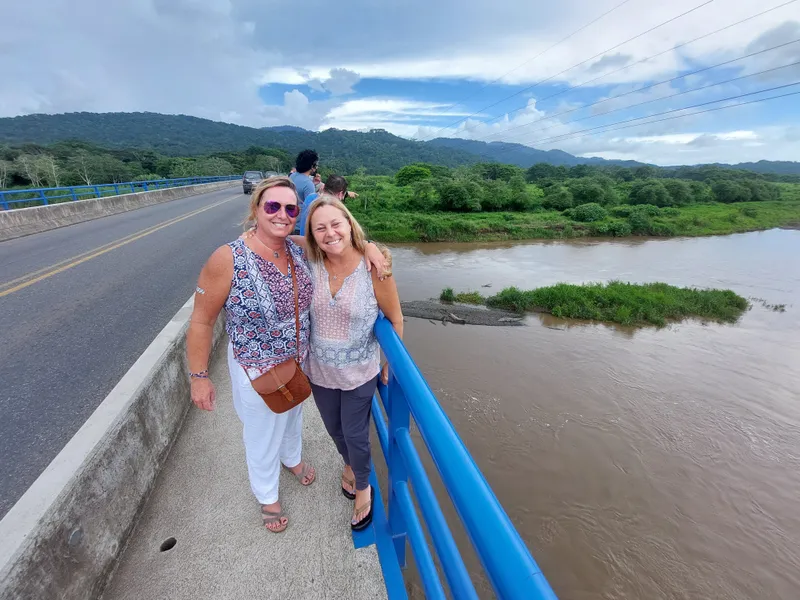 Two women lean on the blue railing of a bridge over a wide brown river, green hills behind.