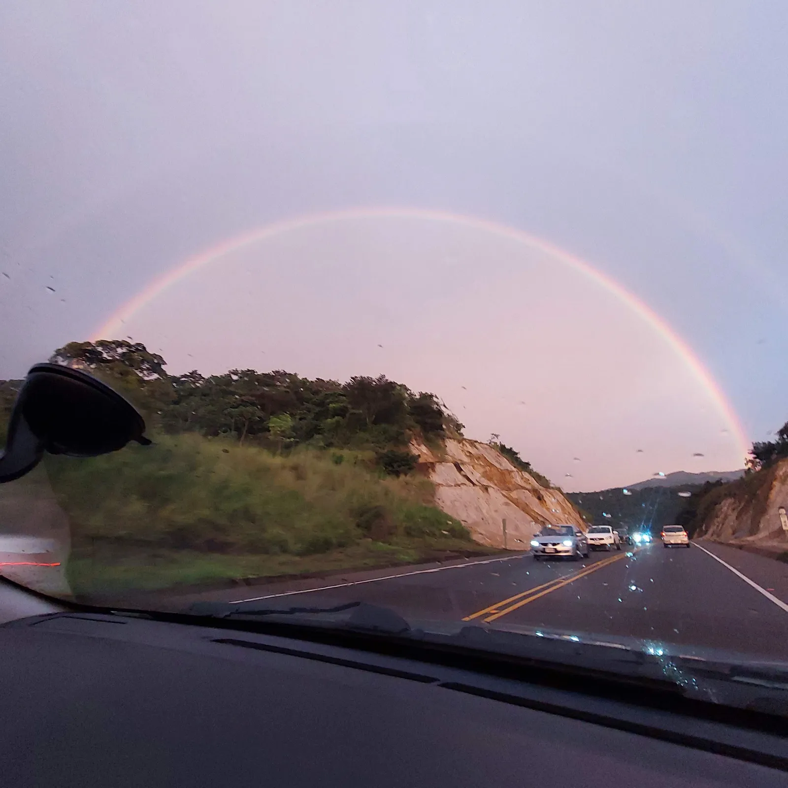 Seen from inside a car, the road drops toward the coast under a full rainbow.