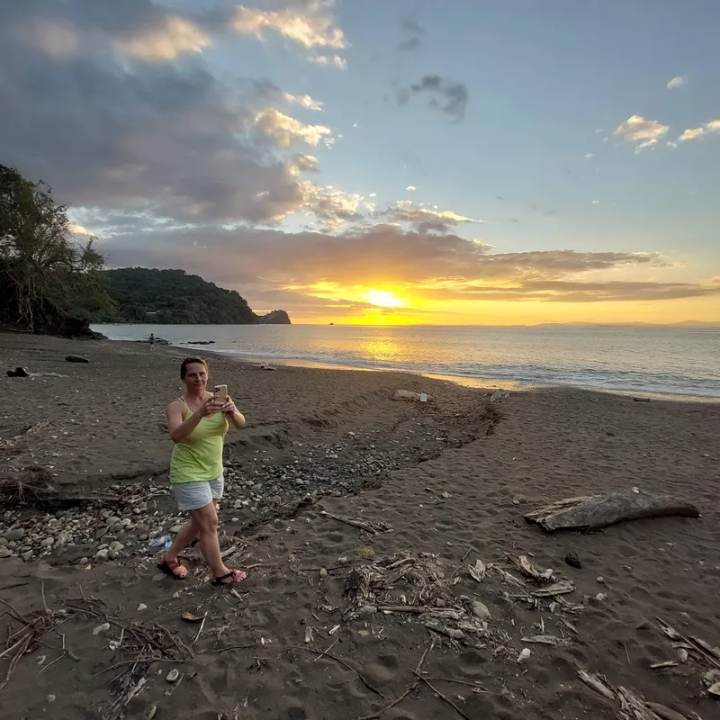 A woman photographs the sunset from a beach scattered with driftwood.