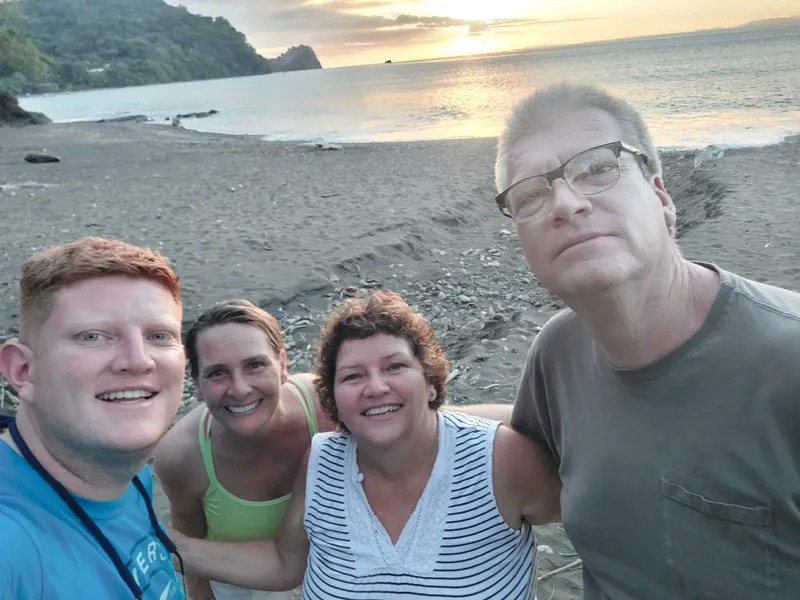 Four people crowd into a selfie on a grey sand beach at dusk.