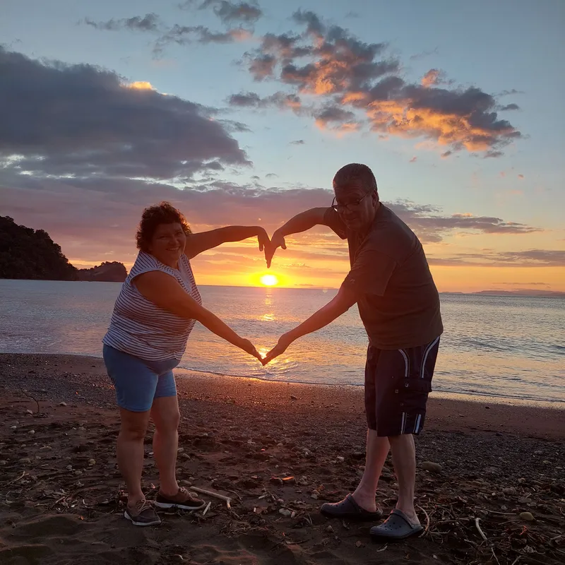 Two people join their arms into a heart shape around the setting sun.