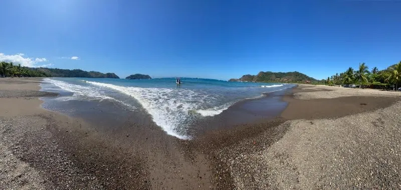 Foam runs up dark sand, with a green headland and a rocky islet in the distance.