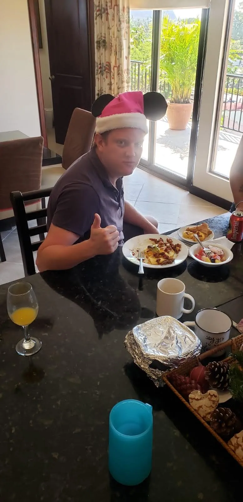 A young man gives a thumbs up at a breakfast table set with plates, fruit and juice.