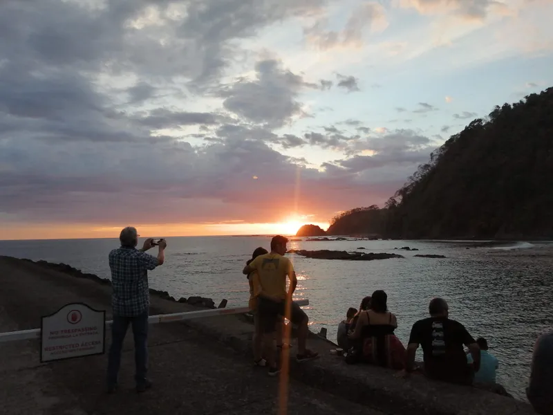 People stand and sit on a lookout, watching the sun set over a rocky cove.