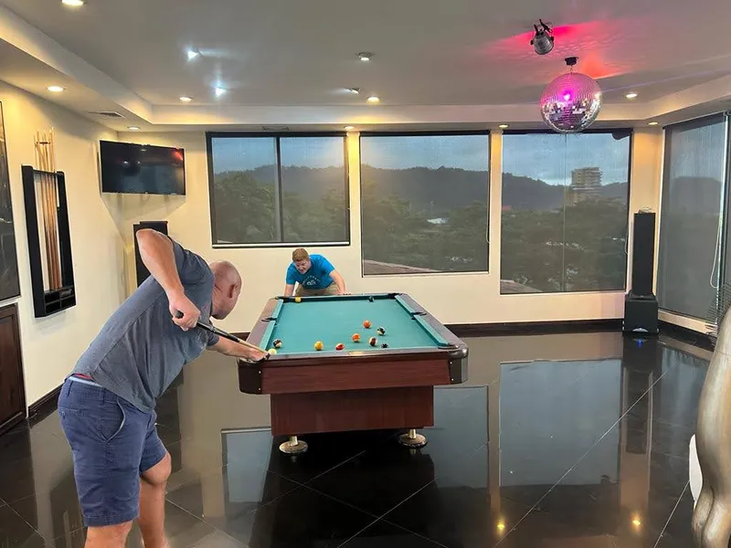 Two people play pool in a room with wide windows onto the hills and a mirror ball overhead.