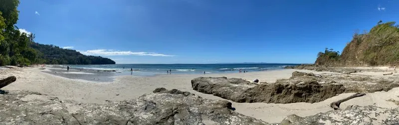 Dark rocks sit on pale sand, with swimmers out in the blue water beyond.