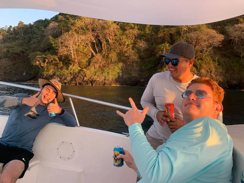Three men with drinks on the deck of a boat, a forested shore lit gold behind them.