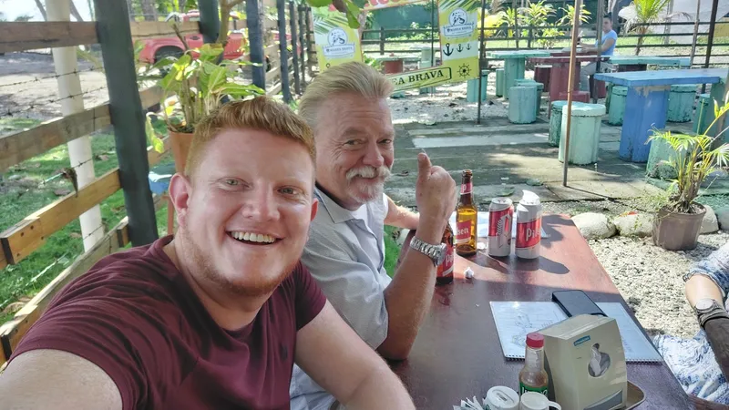 Two men smile at an open air bar table with beers and cans in front of them.