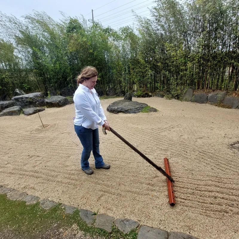 A woman rakes lines into the gravel of a garden, with bamboo growing behind her.