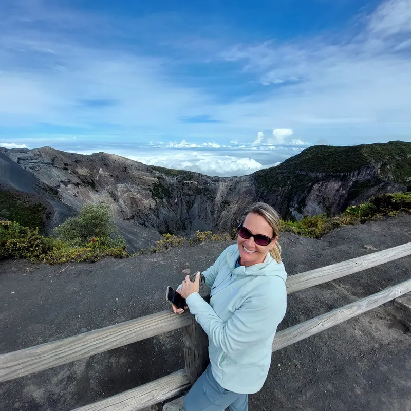 A woman in a light blue sweatshirt leans on a wooden railing at a lookout above a volcanic crater.