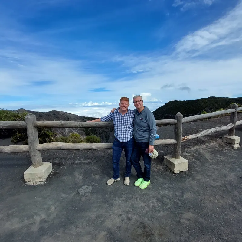 Two men pose side by side at a wooden railing, with dark ridges and open sky behind them.