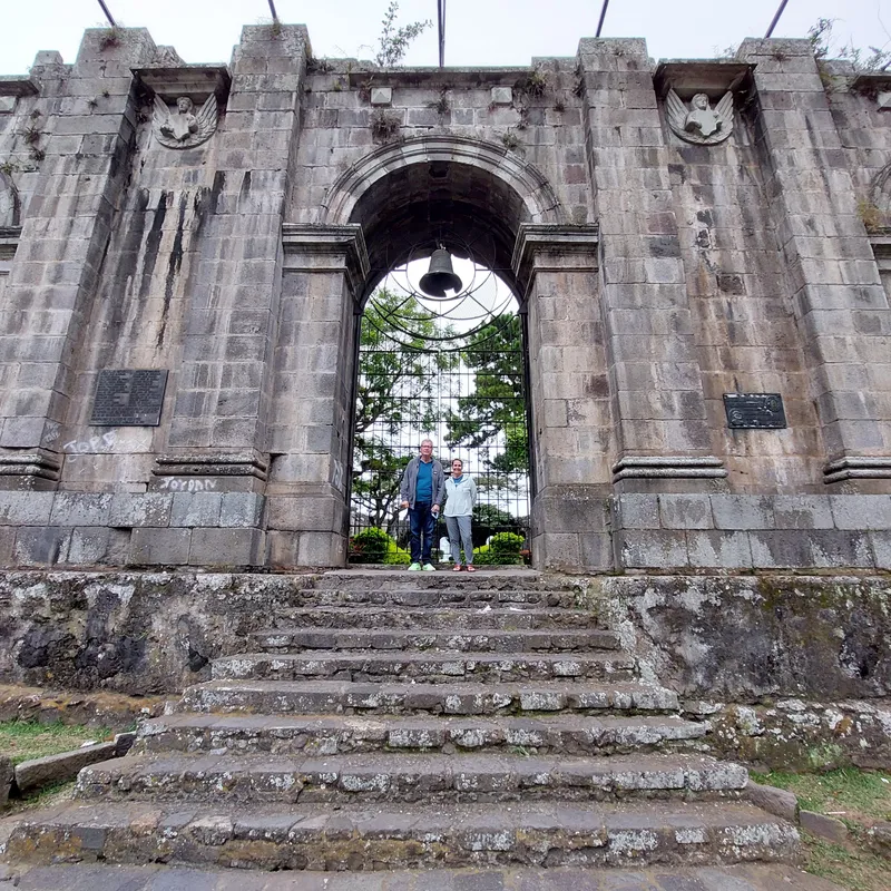 Stone church ruins with a bell hanging in the central arch and a couple standing at the top of the steps.