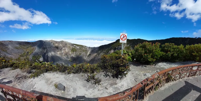 A wide view of a crater from a gravel lookout, with a sign warning against going past the edge.