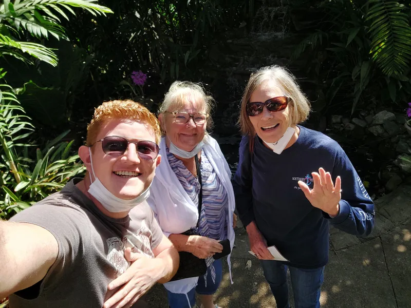 Three people take a selfie in a garden, masks pulled down and one of them waving.