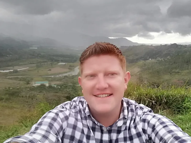 A man takes a selfie on a hilltop, with a green valley and a rainy sky behind him.