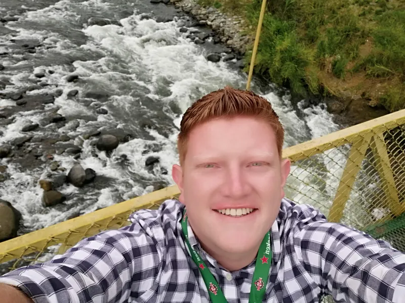 A man takes a selfie on a bridge with a yellow railing, over a river running white between rocks.