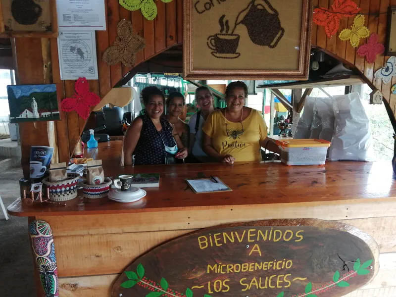Four women behind the wooden counter of a coffee mill shop, with a picture made of coffee beans on the wall and a painted welcome sign in front.