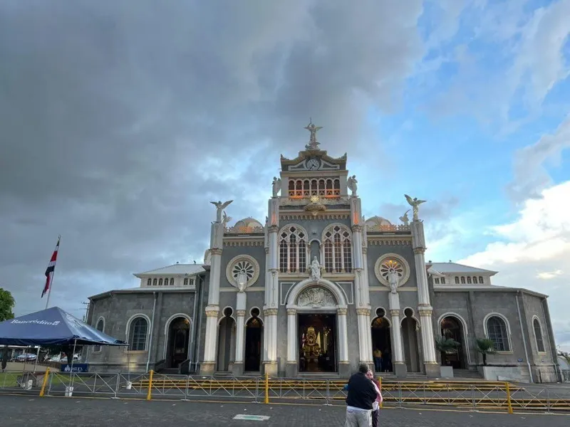 The front of a large church with angel statues under a heavy grey sky, one person standing in the plaza.