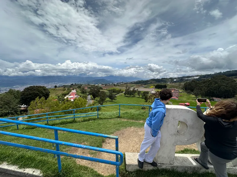A boy in a blue sweatshirt leans against a large concrete letter at a lookout, with blue railings and a valley of fields behind.