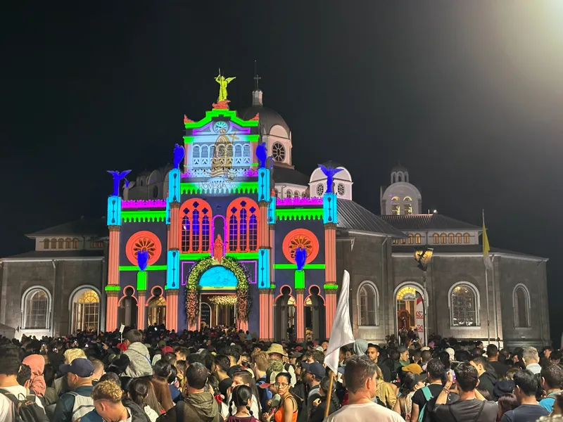 A church front lit with colored projections at night, a crowd filling the plaza in front of it.