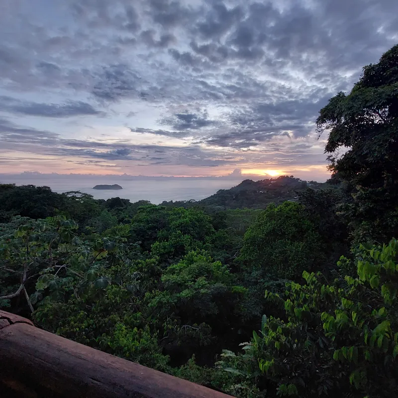 Sunset over forested hills, with the sea and a small island on the horizon under heavy clouds.