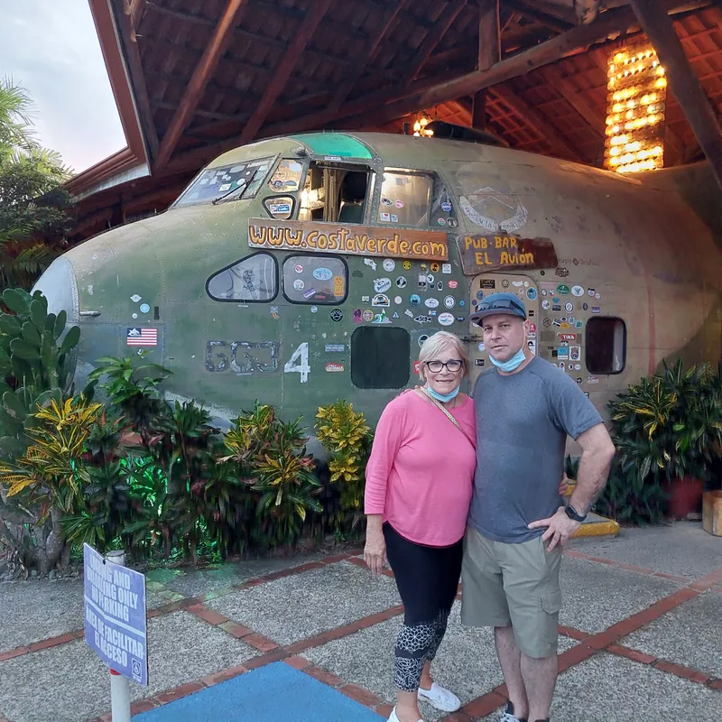 A couple poses in front of the green fuselage of an old plane covered in stickers, under a tall roof.