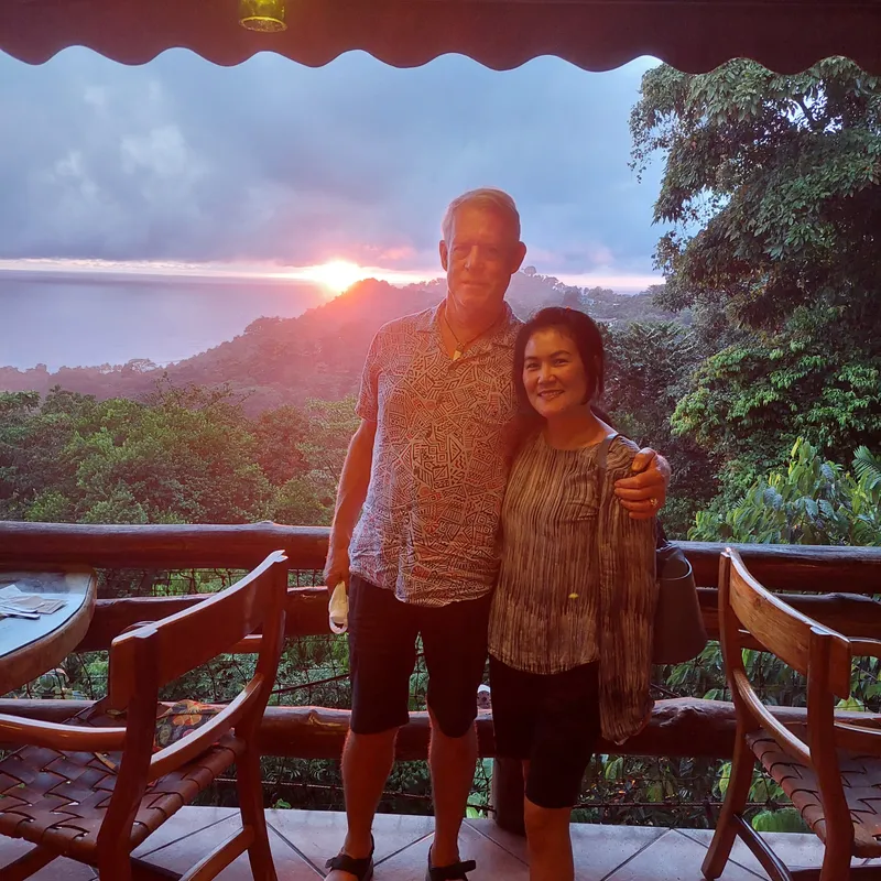 A couple stands on a wooden terrace at sunset, with forest and the last light behind them.