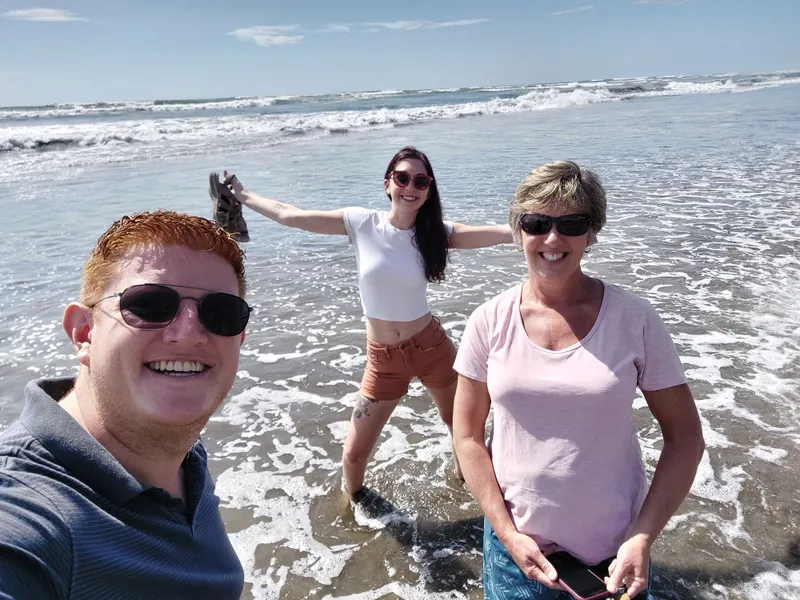 Three people stand in the shallow surf, one of them taking the photo up close.