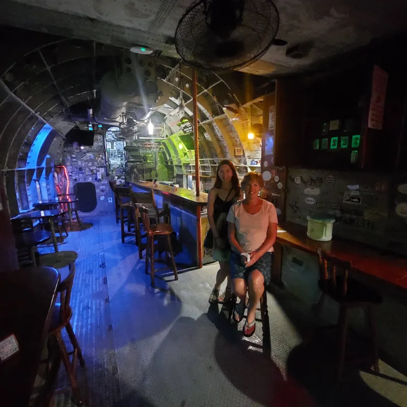 The inside of a bar built into an airplane fuselage, blue light along the curved ceiling and two people sitting at the counter.