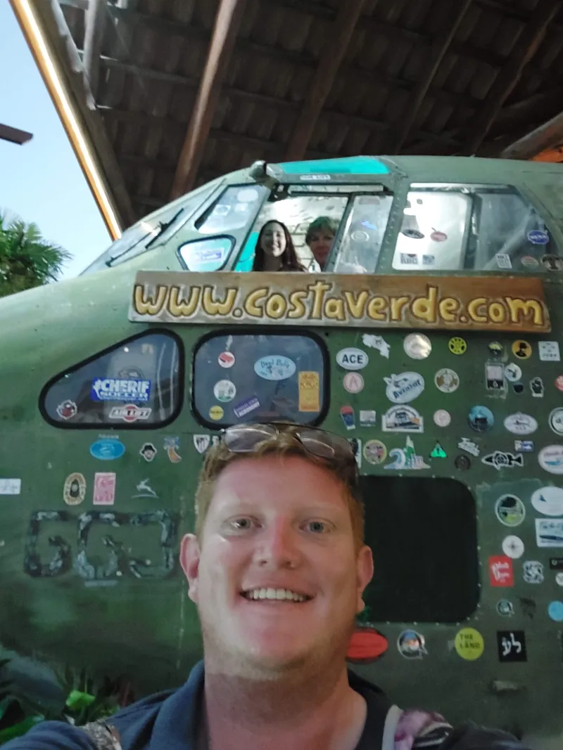 A man takes a selfie in front of the sticker covered nose of an old plane, with other people reflected in the cockpit windows.