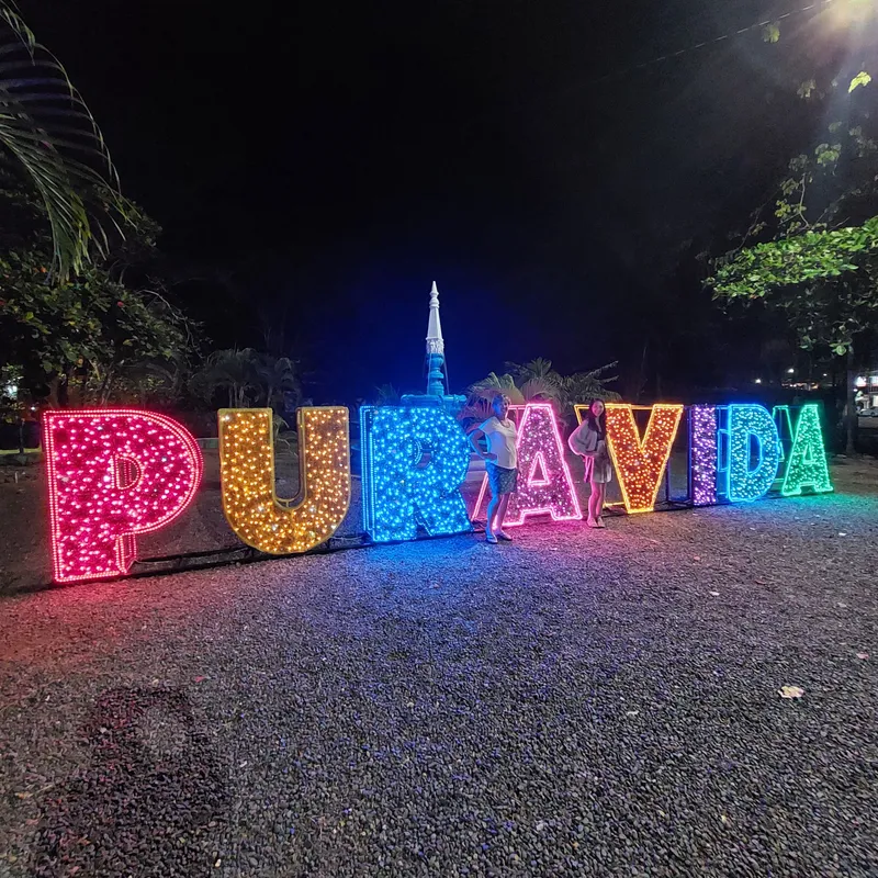 Large lit up letters spelling PURA VIDA glow in different colors at night beside a road.