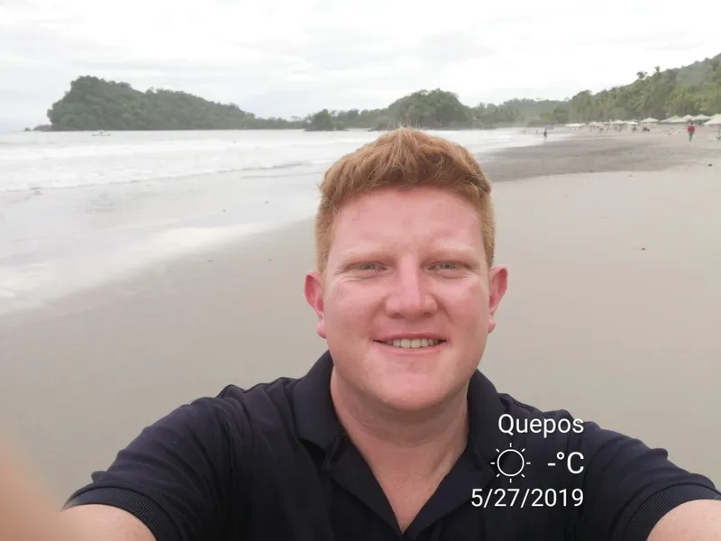 A man takes a selfie on a grey beach, with a forested headland behind him.
