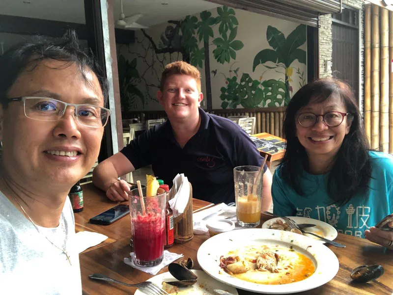Three people smile at a restaurant table set with cold drinks and plates of food.