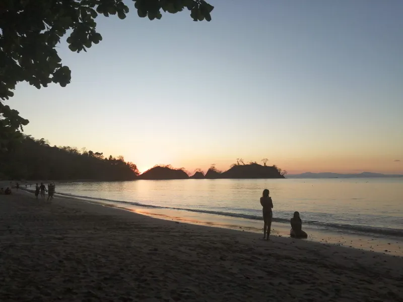 Two figures at the water's edge at sunset, with small islands dark against the sky.